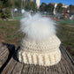 Bonnet bébé écru crocheté à la main, avec revers à reliefs et grand pompon blanc, posé sur un banc en bois