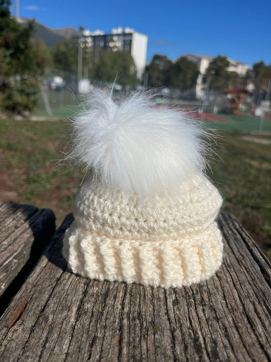 Bonnet bébé écru crocheté à la main, avec revers à reliefs et grand pompon blanc, posé sur un banc en bois